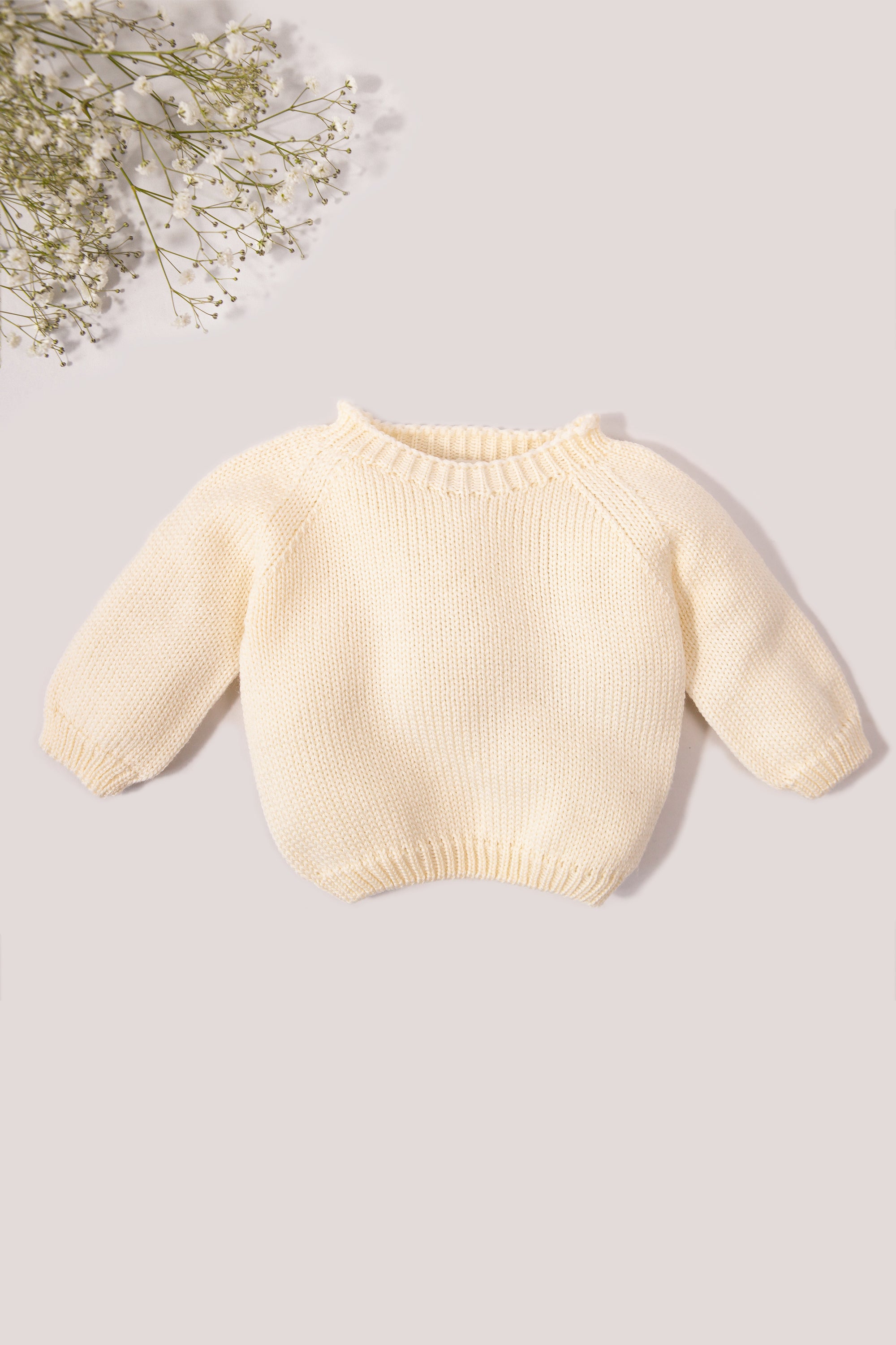 Cream-colored baby sweater on a light gray background with a small bouquet of flowers.